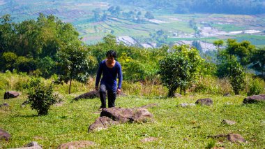 Bir dağ vadisinde genç bir adam, Cavalı bir adam vadinin zirvesine doğru uzun bir tişörtle yürüyor, dağlarda yürüyen genç Asyalı bir adam..