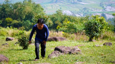 Bir dağ vadisinde genç bir adam, Cavalı bir adam vadinin zirvesine doğru uzun bir tişörtle yürüyor, dağlarda yürüyen genç Asyalı bir adam..