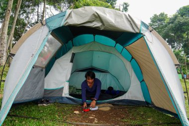 Kamp çadırında oturan mavi gömlekli bir adam çadır kapısının önündeki mini ocağı açmaya çalışıyor. Dağda tatildeyken yemek pişirme kavramı..