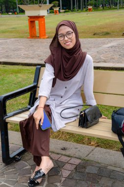 Parkta otururken kameraya gülümseyen, gözlüklü, tesettürlü güzel Asyalı bir kızın ön görüntüsü..