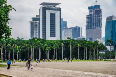 Merkez jakarta, 25 Aralık 2024, bahçede tatil yapan bir ailenin uzak görüntüsü, gökdelenlerin arka planına paralel büyüyen ağaçlar ve boş reklam alanı.