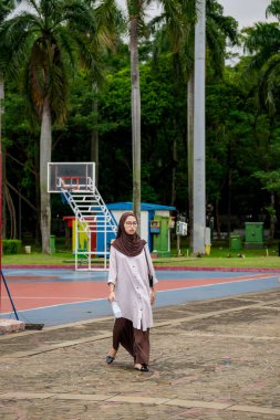 Gözlüklü, omuzunda küçük bir çantayla parkta yürüyen Müslüman bir kadının dikey görüntüsü..