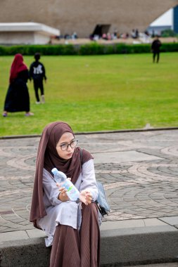 Gözlüklü Müslüman bir kadın yol kenarında oturmuş elinde bir şişe içme suyu tutuyor..