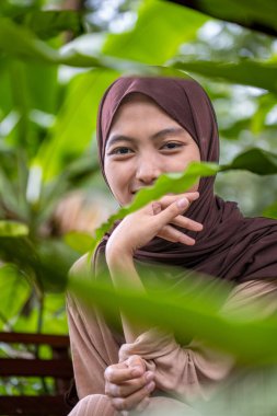Genç müslüman kadın kameraya gülümsüyor çerçevenin arasında yapraklarla. Yeşil bitkilerle süslenmiş genç bir müslüman kadının portresi..