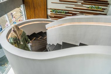 Modern binada dairesel şekilli merdivenler ahşap arka planda ve duvarda süs bitkileri olan modern bir otelin iç mimarisi, modern bir bina iç mimarisi tesettürlü bir kadın aşağı iniyor.