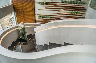Modern binada dairesel şekilli merdivenler ahşap arka planda ve duvarda süs bitkileri olan modern bir otelin iç mimarisi, modern bir bina iç mimarisi tesettürlü bir kadın aşağı iniyor.