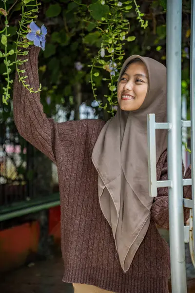 Tesettürdeki kadın elinde hala ağaçta asılı duran bir Madia çiçeği (thunbergia grandiflora) tutuyor.,