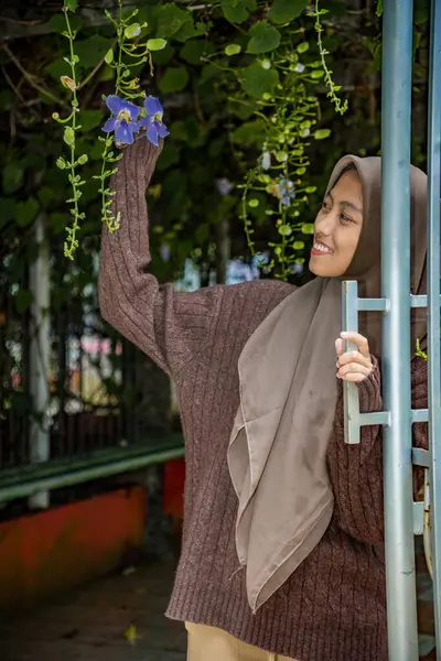 Tesettürdeki kadın elinde hala ağaçta asılı duran bir Madia çiçeği (thunbergia grandiflora) tutuyor.,