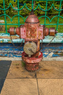 Arka planda demir bir çitle kaldırımda paslı bir yangın söndürücünün görüntüsü.