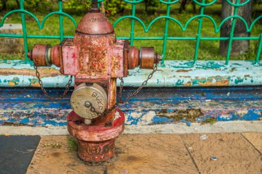 Arka planda demir bir çitle kaldırımda paslı bir yangın söndürücünün görüntüsü.