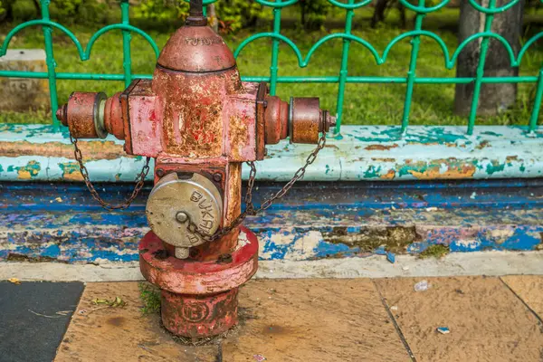 Arka planda demir bir çitle kaldırımda paslı bir yangın söndürücünün görüntüsü.