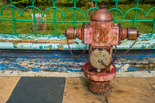 Arka planda demir bir çitle kaldırımda paslı bir yangın söndürücünün görüntüsü.