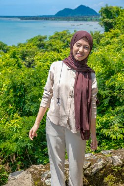 Kayalıklarda duran genç bir kadın plaj arka planında, güzel bir Asyalı kadın günlük kıyafetler giyiyor, gökyüzü ile birlikte boş bir uçurumda poz veriyor, moda fotoğrafı konsepti..