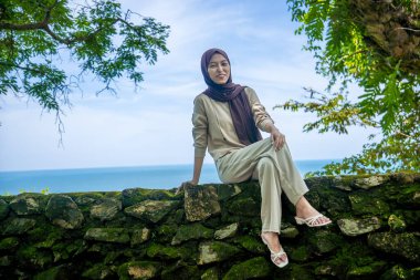 Kayalıklarda, plaj arka planında oturan genç bir kadın, sıradan kıyafetler giyen güzel bir Asyalı kadın, boş bir yer olarak gökyüzünde poz veren bir uçurum, moda fotoğrafı konsepti..