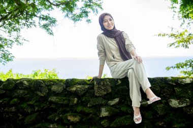 Kayalıklarda, plaj arka planında oturan genç bir kadın, sıradan kıyafetler giyen güzel bir Asyalı kadın, boş bir yer olarak gökyüzünde poz veren bir uçurum, moda fotoğrafı konsepti..