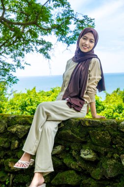 Kayalıklarda, plaj arka planında oturan genç bir kadın, sıradan kıyafetler giyen güzel bir Asyalı kadın, boş bir yer olarak gökyüzünde poz veren bir uçurum, moda fotoğrafı konsepti..