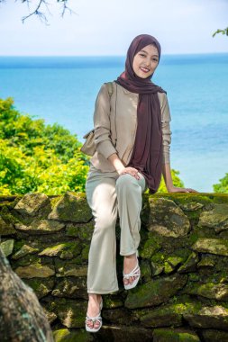 Kayalıklarda, plaj arka planında oturan genç bir kadın, sıradan kıyafetler giyen güzel bir Asyalı kadın, boş bir yer olarak gökyüzünde poz veren bir uçurum, moda fotoğrafı konsepti..