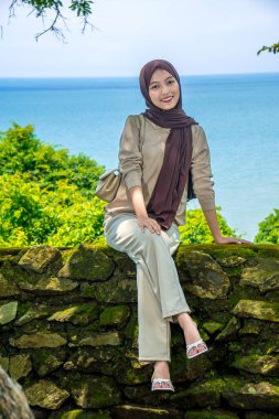 Kayalıklarda, plaj arka planında oturan genç bir kadın, sıradan kıyafetler giyen güzel bir Asyalı kadın, boş bir yer olarak gökyüzünde poz veren bir uçurum, moda fotoğrafı konsepti..