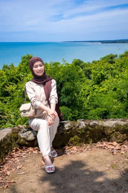 Kayalıklarda, plaj arka planında oturan genç bir kadın, sıradan kıyafetler giyen güzel bir Asyalı kadın, boş bir yer olarak gökyüzünde poz veren bir uçurum, moda fotoğrafı konsepti..