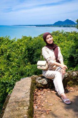 Kayalıklarda, plaj arka planında oturan genç bir kadın, sıradan kıyafetler giyen güzel bir Asyalı kadın, boş bir yer olarak gökyüzünde poz veren bir uçurum, moda fotoğrafı konsepti..