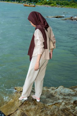 Başörtüsü takan Müslüman kız, sahilde kayalıklarda ayaklarını oynayarak rahatlıyor. Müslüman kadın sahilde kayalıklarda ayaklarıyla oynuyor..