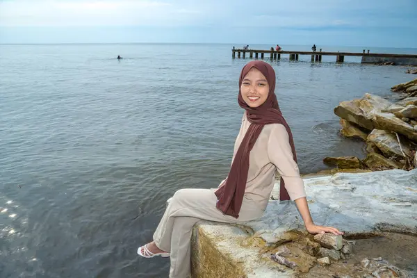 Asyalı Müslüman kadın deniz kenarındaki bir kayanın üzerinde oturuyor. Arka planda sakin bir deniz suyu ve gökyüzü reklam için boş bir alan. Deniz kenarındaki bir kayanın üzerinde oturan kadın kameraya bakıyor..
