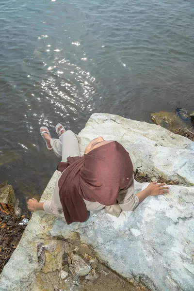 Asyalı Müslüman bir kızın deniz kenarındaki bir kayanın üzerinde oturmuş atmosferin tadını çıkarırken görüntüsü. Dinlenme konsepti, rahatla, mola ver.