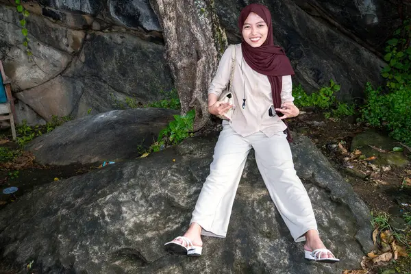 Müslüman Asyalı kadınlar, yosunlu kayalardan oluşan bir ağacın yanındaki büyük bir kayanın üzerinde küçük bir çanta taşıyorlar..