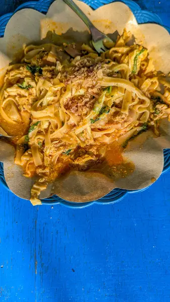 Üzerine kızarmış soğan serpiştirilmiş kalın kahverengi soslu bir porsiyon erişte kaşık ve çatalla kaplanmış kağıt tabakta servis edilir..