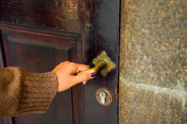 Süveter giyen bir kadının eli eski ahşap bir kapı tokmağını tutuyor..