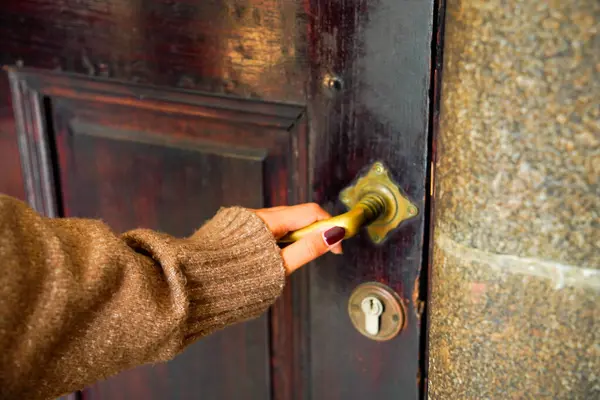 Süveter giyen bir kadının eli eski ahşap bir kapı tokmağını tutuyor..