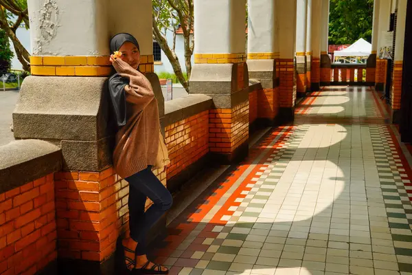 Başörtülü bir kadın, uzun bir koridor ve yerde dramatik gölgeler bırakan güneş ışığı olan bir binada kırmızı tuğlalı bir sütunun yanında duruyor..