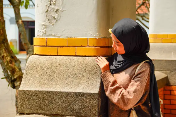 Başörtülü bir kadın, uzun bir koridor ve yerde dramatik gölgeler bırakan güneş ışığı olan bir binada kırmızı tuğlalı bir sütunun yanında duruyor..