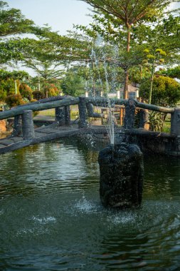 Huzurlu bir göletin üzerindeki kemerli taş köprü yemyeşil ağaçlarla çevrili bir çeşme ve yemyeşil bir bahçedeki çocuk oyun alanı..