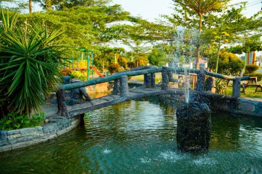Huzurlu bir göletin üzerindeki kemerli taş köprü yemyeşil ağaçlarla çevrili bir çeşme ve yemyeşil bir bahçedeki çocuk oyun alanı..