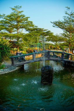 Huzurlu bir göletin üzerindeki kemerli taş köprü yemyeşil ağaçlarla çevrili bir çeşme ve yemyeşil bir bahçedeki çocuk oyun alanı..