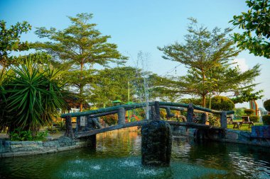 Huzurlu bir göletin üzerindeki kemerli taş köprü yemyeşil ağaçlarla çevrili bir çeşme ve yemyeşil bir bahçedeki çocuk oyun alanı..
