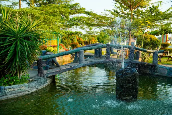Huzurlu bir göletin üzerindeki kemerli taş köprü yemyeşil ağaçlarla çevrili bir çeşme ve yemyeşil bir bahçedeki çocuk oyun alanı..