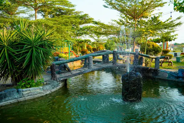 Huzurlu bir göletin üzerindeki kemerli taş köprü yemyeşil ağaçlarla çevrili bir çeşme ve yemyeşil bir bahçedeki çocuk oyun alanı..