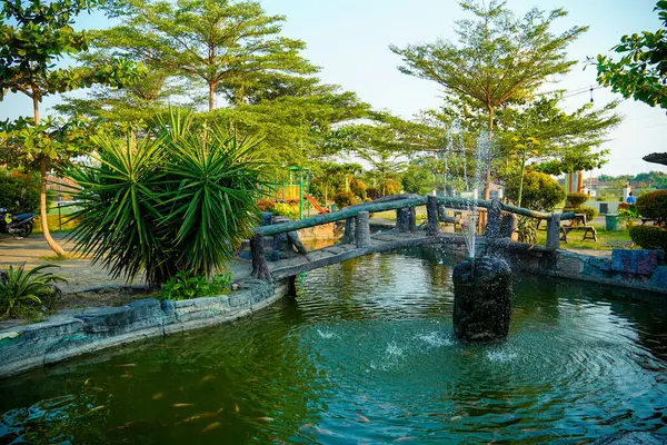 Huzurlu bir göletin üzerindeki kemerli taş köprü yemyeşil ağaçlarla çevrili bir çeşme ve yemyeşil bir bahçedeki çocuk oyun alanı..