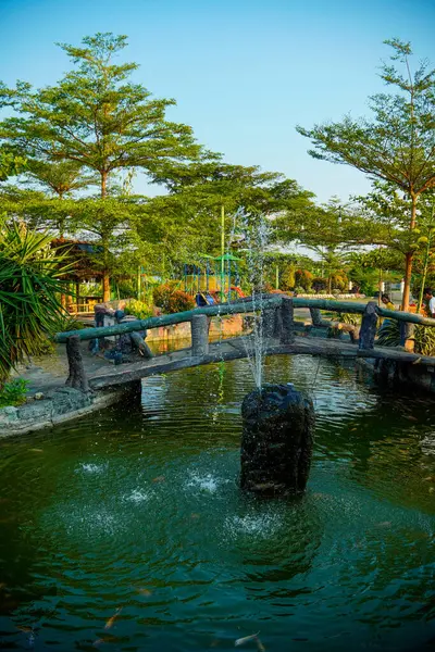 Huzurlu bir göletin üzerindeki kemerli taş köprü yemyeşil ağaçlarla çevrili bir çeşme ve yemyeşil bir bahçedeki çocuk oyun alanı..