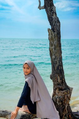 Bir kadın tesettürlü bir ağaç gövdesinde oturuyor kumlu bir plajın kenarında, sakin bir deniz dalgası ve reklam alanı için açık bir gökyüzü ile.,