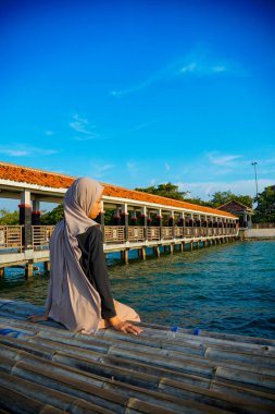 Başörtülü bir kadın, arkasında kırmızı çatılı bir iskele ve reklam alanı için arka planda parlak bir gökyüzü olan sakin bir denizin kenarında bambunun üzerine oturur..