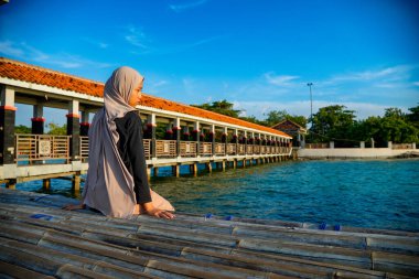 Başörtülü bir kadın, arkasında kırmızı çatılı bir iskele ve reklam alanı için arka planda parlak bir gökyüzü olan sakin bir denizin kenarında bambunun üzerine oturur..