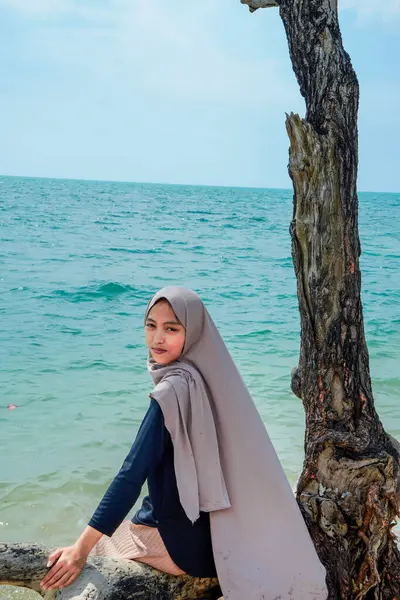 Bir kadın tesettürlü bir ağaç gövdesinde oturuyor kumlu bir plajın kenarında, sakin bir deniz dalgası ve reklam alanı için açık bir gökyüzü ile.,