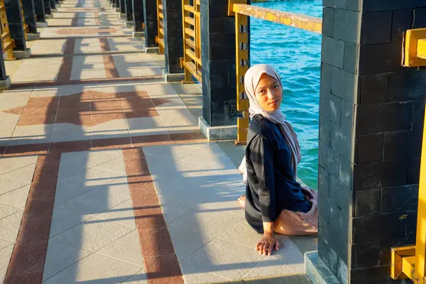 Asyalı Müslüman kadın köprü sütunlarından gölgeler taşıyan yayalar için denizin üzerinde uzun bir köprüde oturuyor..