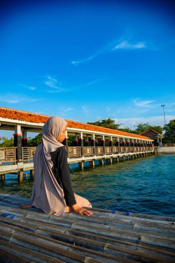Başörtülü bir kadın, arkasında kırmızı çatılı bir iskele ve reklam alanı için arka planda parlak bir gökyüzü olan sakin bir denizin kenarında bambunun üzerine oturur..