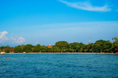 Sahilde ağaçların olduğu sığ bir kumsalda küçük dalgalar kırılıyor ve reklam alanı için açık bir gökyüzü var..