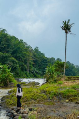 Dağ manzarası, küçük şelaleden akan nehirler ve önplanda duran kadın, tesetteki kayaların üzerinde akan kayaların yanında duran tesettür ve berrak gökyüzü.
