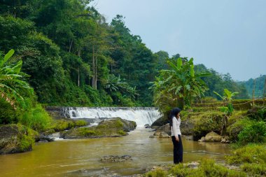 Dağ manzarası, küçük şelaleden akan nehirler ve önplanda duran kadın, tesetteki kayaların üzerinde akan kayaların yanında duran tesettür ve berrak gökyüzü.